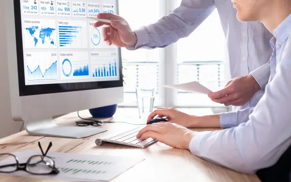 Photograph of two people analyzing a computer screen displaying various business analytics dashboards, including maps, bar charts, line graphs, and pie charts in blue and white colors