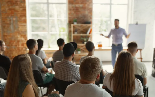 Photograph of a group of people seated and attentively listening to a presenter standing near a flip chart in a bright room with large windows and exposed brick walls. 