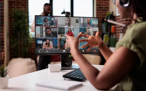 Photograph of a virtual meeting with multiple participants displayed on a computer monitor, showing a grid layout of seven people engaged in discussion. 