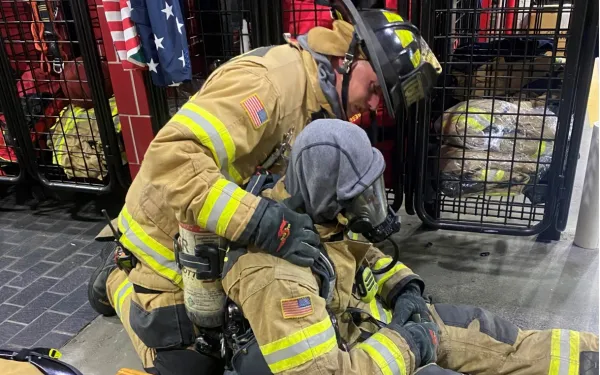 Photograph of two firefighters in full gear inside a fire station, with one kneeling and supporting the other who is seated on the floor wearing a breathing mask. 