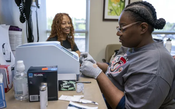 A healthcare worker in gloves uses medical equipment at a desk, while a woman with curly hair smiles in the background. The setting is a clinic room.