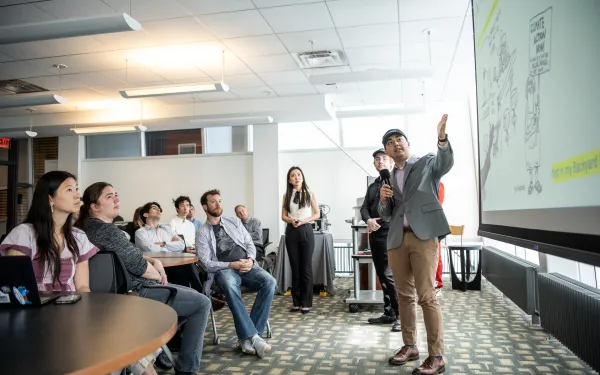 Photograph of a business presentation in a modern office setting, showing a presenter pointing at a projected slide while speaking to an attentive audience seated around tables. 