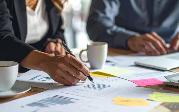 Photograph of two people working at a table covered with printed charts, graphs, and colorful sticky notes, indicating a collaborative business or project planning session.